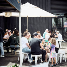 Load image into Gallery viewer, People sitting outdoors under a white umbrella with a 'Strong Organics' sign in the foreground.