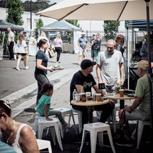 Load image into Gallery viewer, People enjoying outdoor seating at a cafe with tables and umbrellas with market stalls in the background.
