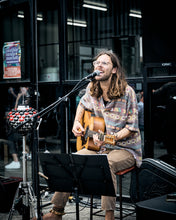Load image into Gallery viewer, Man playing guitar and singing into a microphone in front of a building entrance.
